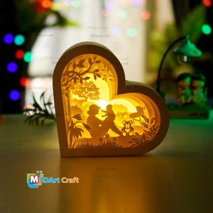 May include: A white wooden heart-shaped lamp with a 3D layered design. The lamp features a silhouette of a parent and child sitting in a forest scene with a teddy bear. The lamp is illuminated with a warm yellow light.