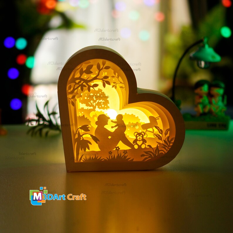 May include: A white wooden heart-shaped lamp with a 3D layered design. The lamp features a silhouette of a parent and child sitting in a forest scene with a teddy bear. The lamp is illuminated with a warm yellow light.