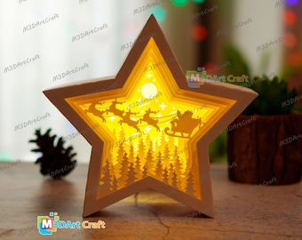 May include: A white star-shaped papercraft with a 3D layered design. The design features a winter scene with a glowing moon, a forest of trees, and Santa Claus in his sleigh pulled by reindeer.