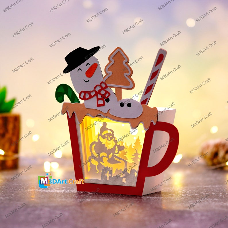 K&ouml;nnte beinhalten: Eine rote und wei&szlig;e Papiercraft-Tasse mit einem Schneemann, einem Weihnachtsbaum und einem Zuckerstangen-Lutscher oben drauf. Die Tasse ist von innen beleuchtet und beleuchtet eine Silhouette des Weihnachtsmanns, der auf einer Bank sitzt.