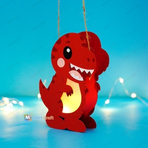 Könnte beinhalten: Eine rote, dinosaurierförmige Papierlaterne in Form eines T-Rex im Cartoon-Stil mit dunkelroten Flecken, einem weißen Bauch und spitzen weißen Zähnen. Die Laterne ist an einer dünnen Schnur aufgehängt und strahlt ein warmes, sanftes Licht aus.