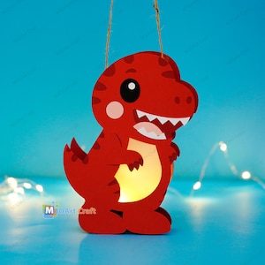 May include: A red dinosaur-shaped hanging lantern with a light-coloured belly, dark red stripes, and a smiling face with white teeth. The lantern is suspended by a thin twine string and features an internal light source that illuminates the belly.