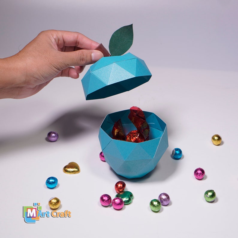 K&ouml;nnte beinhalten: Eine t&uuml;rkisfarbene Origami-Apfelbox mit einem gr&uuml;nen Blatt oben. Die Box ist ge&ouml;ffnet und mit rot und gold folierten Schokoladen gef&uuml;llt. Die Box ist von bunten, in Folie verpackten Schokoladen umgeben.