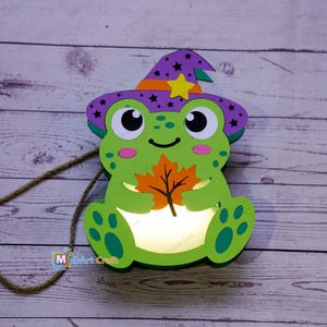 Puede incluir: Una decoración de rana iluminada con cuerpo verde brillante, mejillas rosadas y un sombrero de bruja morado con estrellas negras y una estrella amarilla. La rana sostiene una hoja de arce naranja y el centro del cuerpo emite una luz blanca.