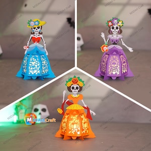 Pode incluir: Três figuras de papel do Dia dos Mortos. Cada figura é um esqueleto vestindo um vestido colorido e uma coroa de flores. Os vestidos são azuis, roxos e laranja. As figuras são rotuladas com "M3D Art Craft".