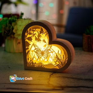 May include: A heart-shaped, illuminated paper cut light box featuring a butterfly and floral design. The light box is made of layered paper, creating a 3D effect. The light source casts a warm glow, highlighting the intricate details of the cutouts. The text "M3dArt Craft" is visible.
