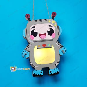 Puede incluir: Una decoración de pared colgante con forma de robot de dibujos animados con cuerpo gris, detalles azules y un panel pectoral amarillo brillante. El robot presenta una cara sonriente, mejillas rosadas, dos antenas con puntas rojas y estrellas amarillas.