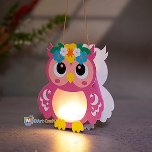 Może przedstawiać: Różowy papierowy lampion w kształcie sowy z wieńcem z niebieskich, żółtych i białych kwiatów, dużymi czarnymi oczami, żółtym dziobem i białymi wzorami półksiężyców na skrzydłach. Środek świeci ciepłym światłem i wisi na cienkim sznurku.