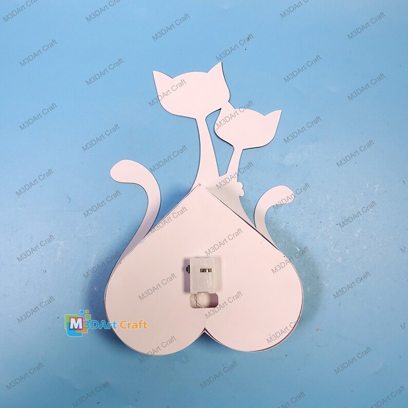 May include: White cutout of two cats in a heart shape. The cats are facing each other and have their tails intertwined. The heart shape has a small light bulb in the center.