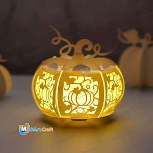Può includere: Una lanterna di carta a forma di zucca gialla con intricati ritagli di foglie e zucche. La lanterna è illuminata dall'interno, creando un bagliore caldo.