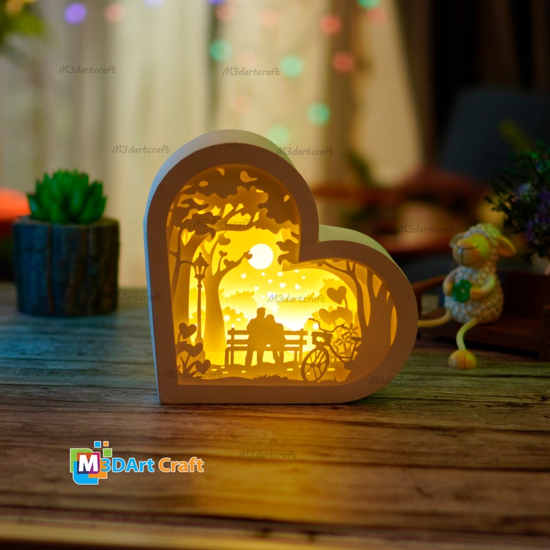 May include: A white wooden heart-shaped lamp with a cutout design of a couple sitting on a bench in a park with a bicycle and a street lamp. The lamp is lit up and casts a warm glow.