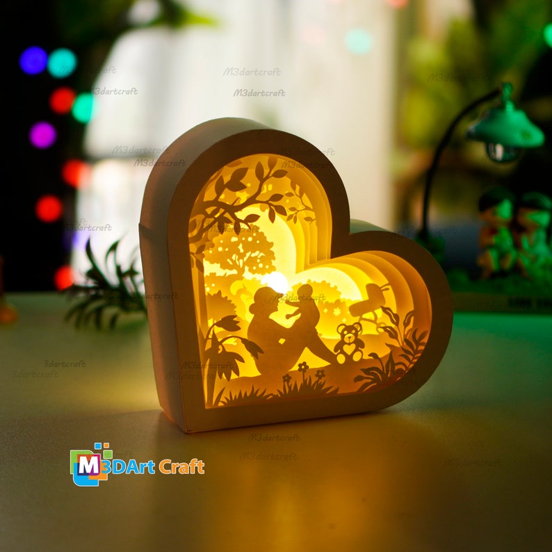 May include: A white heart-shaped 3D papercraft lamp with a cutout design of a mother and child sitting in a forest scene. The lamp is lit up from the inside, casting a warm glow.