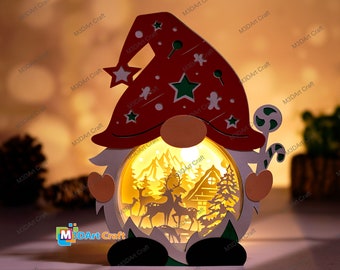Pode incluir: Uma decoração de gnomo de Natal de madeira com luz, com um chapéu vermelho, barba branca e um bastão de caramelo. O gnomo está segurando uma cena iluminada de um país das maravilhas de inverno com uma cabana, árvores e veados.