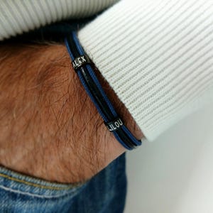 Peut inclure: Un bracelet noir et bleu avec les noms "ALEX" et "LILOU" gravés sur des perles argentées. Le bracelet est porté sur un poignet, avec un pull blanc et un jean bleu en arrière-plan.