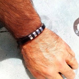 Peut inclure: Un bracelet en cuir noir avec des accents en métal argenté. Le bracelet est porté au poignet.