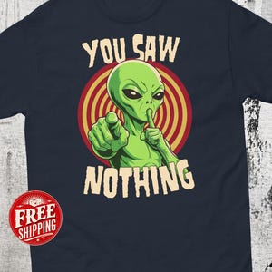 May include: A dark navy blue t-shirt featuring a graphic of a green alien with large black eyes. The alien is pointing forward with one hand and holding a finger to its lips with the other. The text YOU SAW NOTHING is printed in beige.
