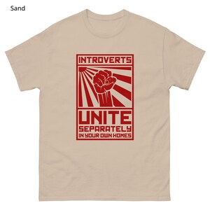 May include: Sand-colored t-shirt with a red graphic. The graphic features the word "INTROVERTS" above a clenched fist, sun rays, and the word "UNITE" with the phrase "SEPARATELY IN YOUR OWN HOMES" below. The shirt is made of a soft material.
