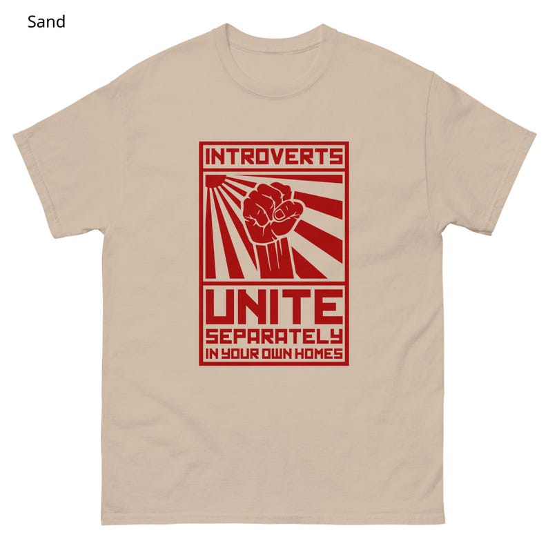 May include: Sand-colored t-shirt with a red graphic. The graphic features the word "INTROVERTS" above a clenched fist, sun rays, and the word "UNITE" with the phrase "SEPARATELY IN YOUR OWN HOMES" below. The shirt is made of a soft material.