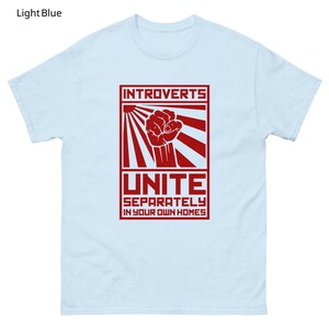 May include: Light blue t-shirt with a red graphic. The graphic features the word "INTROVERTS" above a fist inside a rectangle with sun rays, and the word "UNITE" below, with the text "SEPARATELY IN YOUR OWN HOMES".