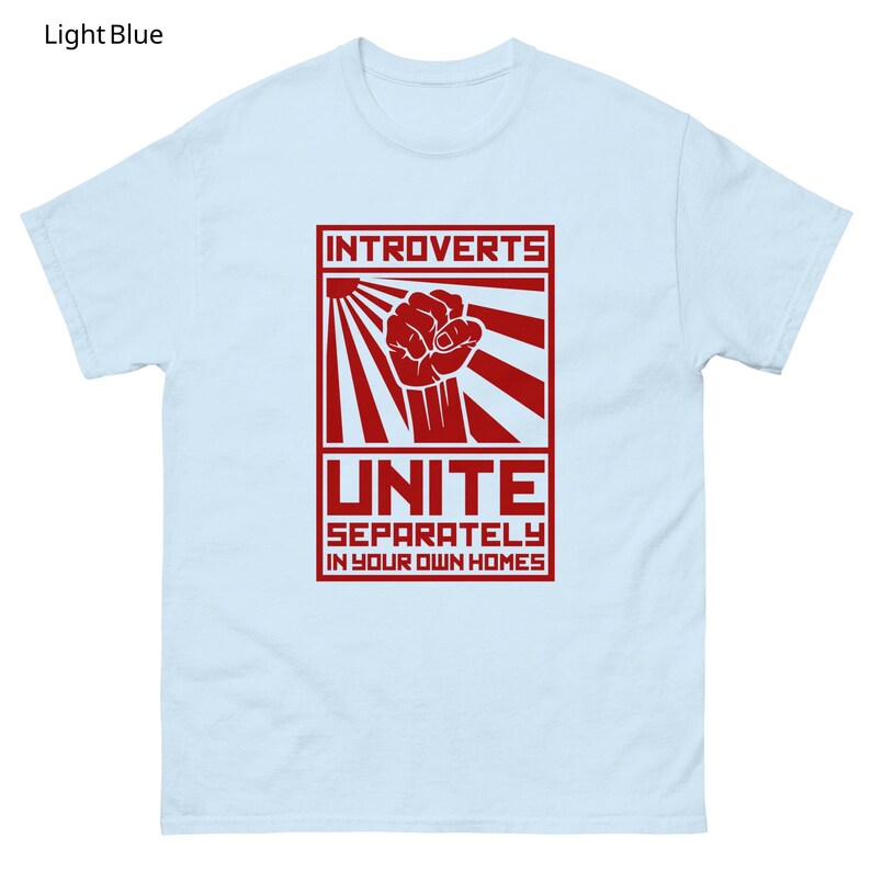 May include: Light blue t-shirt with a red graphic. The graphic features the word "INTROVERTS" above a fist inside a rectangle with sun rays, and the word "UNITE" below, with the text "SEPARATELY IN YOUR OWN HOMES".