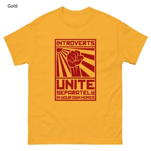 May include: Gold-colored t-shirt with a red graphic. The graphic features the word "INTROVERTS" above a clenched fist and the word "UNITE" with the phrase "SEPARATELY IN YOUR OWN HOMES" below. The shirt is made of a soft material.