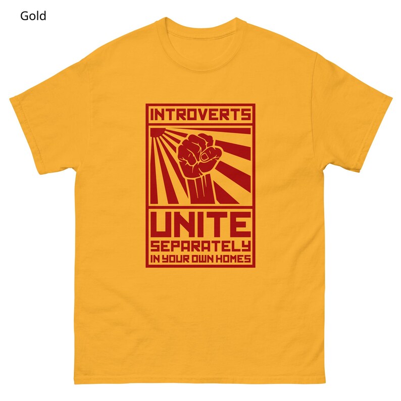May include: Gold-colored t-shirt with a red graphic. The graphic features the word "INTROVERTS" above a clenched fist and the word "UNITE" with the phrase "SEPARATELY IN YOUR OWN HOMES" below. The shirt is made of a soft material.