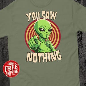 Könnte beinhalten: Ein olivgrünes T-Shirt mit einer Grafik eines grünen Außerirdischen, der mit einer Hand nach vorne zeigt und den Finger der anderen Hand auf die Lippen legt. Dahinter ist ein rot-gelbes Kreismuster. Der Text lautet YOU SAW NOTHING.