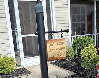 May include: A black metal lamp post with a decorative lantern top and a wooden sign. The sign reads "John - Karen Steven & Baldwin" with a baby graphic. The lamp post is in front of a house.