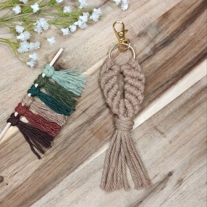 Puede incluir: Un llavero de macramé beige con un anillo de llave dorado y un borla. El llavero está hecho con una cuerda anudada y tiene un diseño en forma de corazón.