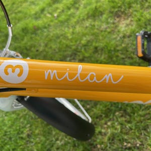 Könnte beinhalten: Ein gelber Fahrradrahmen mit einem weißen Aufkleber, auf dem "milan" steht, und einem weißen Kreis mit der Zahl "3" darin.