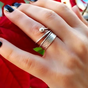 May include: A hand wearing three silver rings. The rings include a hammered band, a spiral ring, and a ring with a small ball. The background is a blurred red and green.