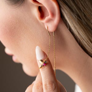 Puede incluir: Pendientes de colibrí dorados con detalles en rubí y negro. Los pendientes tienen una delicada cadena que cuelga de la oreja. El colibrí tiene alas detalladas y un pequeño pico. Los pendientes se están usando.