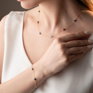 Può includere: Una collana e un braccialetto d'oro con perline quadrate blu e nere. La collana è indossata da una persona con una camicetta bianca.