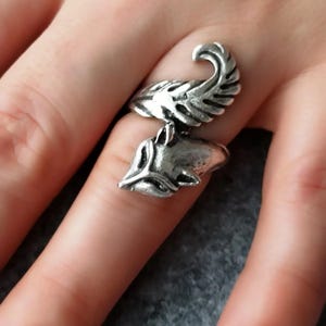 May include: A silver-toned ring featuring a stylized wolf head and feather design. The ring is worn on a finger, showcasing the intricate details of the metalwork. The ring's design is a unique piece of jewelry.