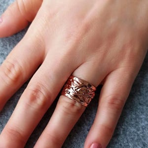 May include: A rose gold ring with an intricate floral design. The wide band ring is worn on a finger, showcasing the detailed relief pattern. The ring's warm color contrasts with the skin tone and the background.