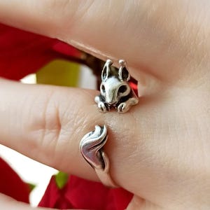 May include: A silver ring with a squirrel design. The squirrel's head and paws are at the top, with its tail forming the ring's band. The ring wraps around the finger. A whimsical piece of jewellery, perfect for animal lovers.