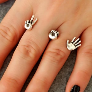 May include: Silver-toned rings featuring hand gestures: a peace sign, a fist, and an open palm. The rings are displayed on a hand with a dark nail polish. The jewelry is likely made of metal and has a detailed, three-dimensional design.
