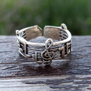 May include: Silver-toned adjustable ring featuring a musical staff design with a treble clef and musical notes. The ring is set against a blurred green and brown background, showcasing the intricate details of the jewelry.