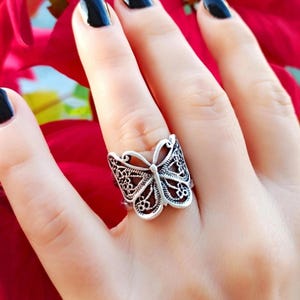 May include: A silver butterfly ring with detailed filigree patterns. The ring showcases openwork wings and a detailed body, creating a delicate and elegant accessory. The ring is shown on a finger against a red background.