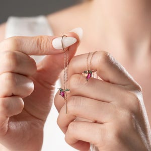 May include: A delicate gold necklace with a hummingbird pendant featuring red and green gemstones.