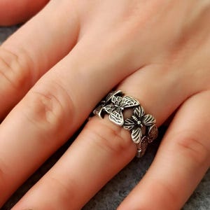 May include: A silver ring with a design of several butterflies. The ring is worn on a finger, highlighting the detailed butterfly shapes. The butterflies have intricate wing patterns and are arranged around the ring's band.