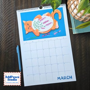 May include: A wall calendar for the month of March featuring an illustration of an orange cat holding an embroidery hoop with lavender sprigs. The calendar grid is white with blue text and lines. A light blue pen is placed next to the calendar.