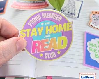 May include: A circular sticker with a purple border and a light blue center. The text reads PROUD MEMBER OF THE STAY HOME AND READ CLUB in yellow, white, and pink lettering, accented by two small yellow stars at the bottom.