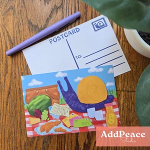 May include: A postcard featuring a snail picnic illustration and a blank postcard with the word "POSTCARD" printed on it. A purple pen lies on the postcards. The snail illustration includes a picnic basket, watermelon, and letters.