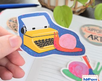 May include: A die-cut sticker featuring a bright yellow typewriter with black keys and a white sheet of paper, positioned next to a pink-shelled snail with a reddish-orange body, all set against a dark blue background with purple speckles.