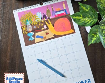 May include: A wall calendar for October featuring an illustration of a yellow snail, a blue oil lamp, crystals, and books on a shelf. The calendar grid is white with blue lines and text, and a blue pen rests on the page. The AddPeace Studio logo is visible.
