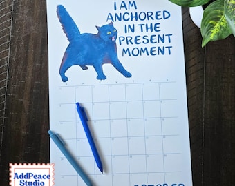 May include: A wall calendar for October featuring a blue cat illustration and the text I AM ANCHORED IN THE PRESENT MOMENT. The calendar grid includes days of the week and dates. Two blue pens are placed diagonally across the calendar page.