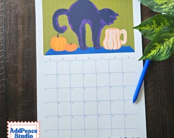 May include: A wall calendar for October featuring a graphic illustration of a dark purple arched-back cat, two orange pumpkins, and a light pink mug on a blue surface against a green chevron background. The calendar grid has blue text.