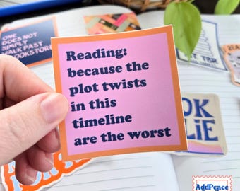May include: A square sticker with a light pink center and an orange border. The text in dark blue reads: Reading: because the plot twists in this timeline are the worst. The sticker is held against a background of a notebook with other stickers.