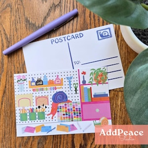May include: A postcard with a blue stamp and address lines, and a decorative postcard featuring a snail and art supplies. A purple pen rests on the postcard. The postcard has the text "POSTCARD".
